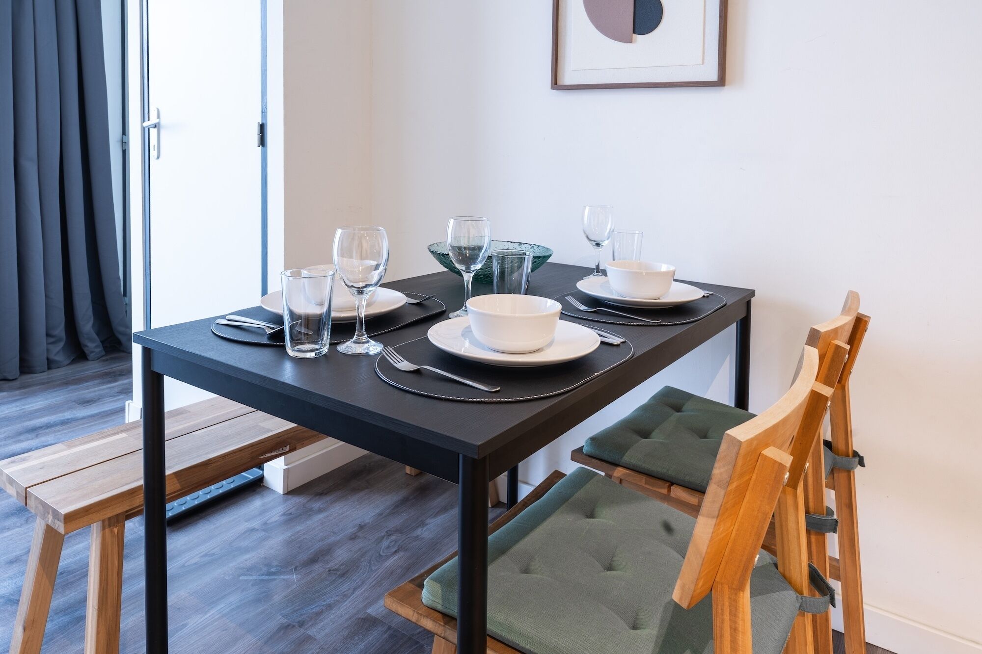 A dining table set with white plates, bowls, and glasses, surrounded by wooden chairs with green cushions.