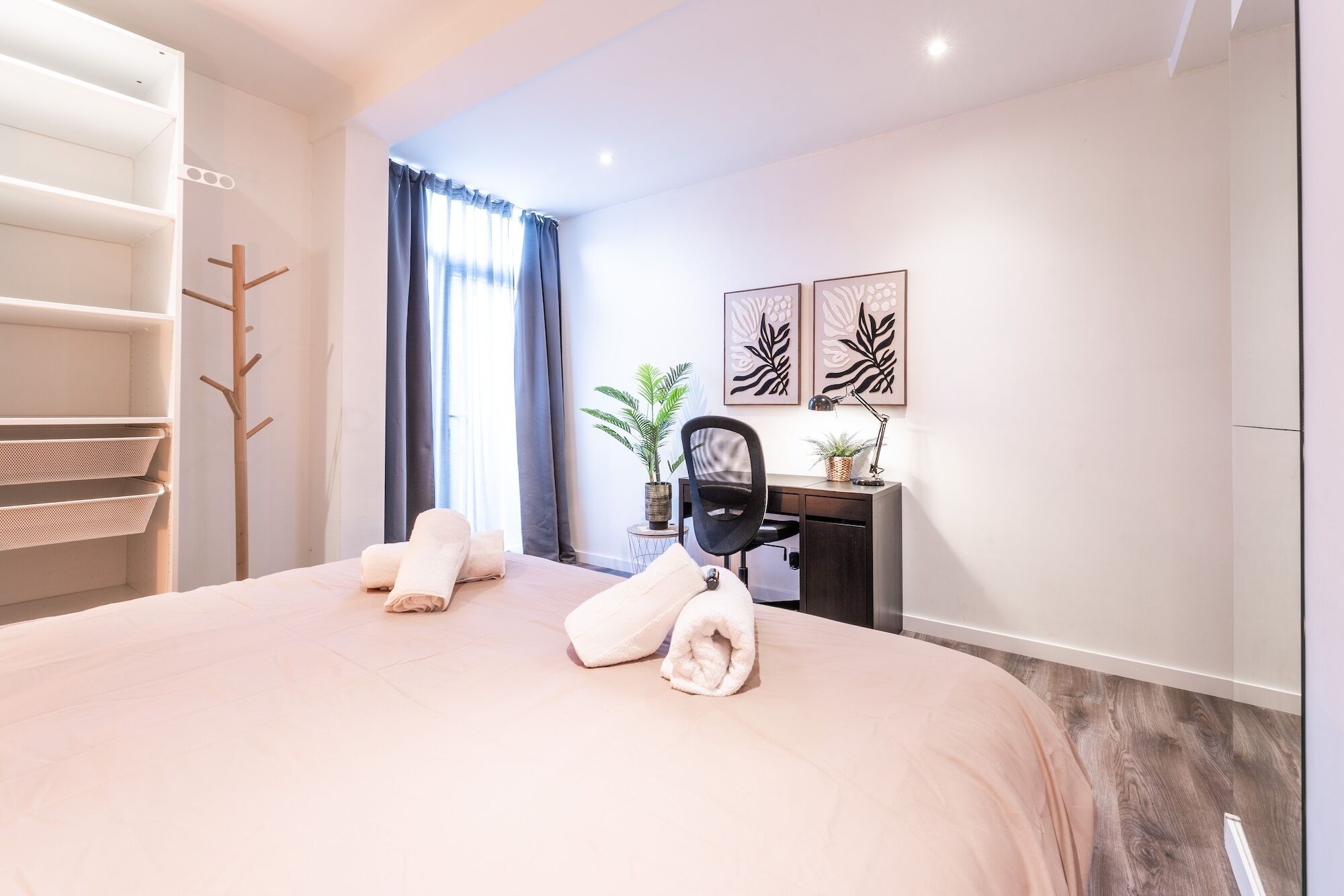 Une chambre d’hôtel comprenant un lit, un bureau, une chaise, un porte-manteau et deux plantes en pot.