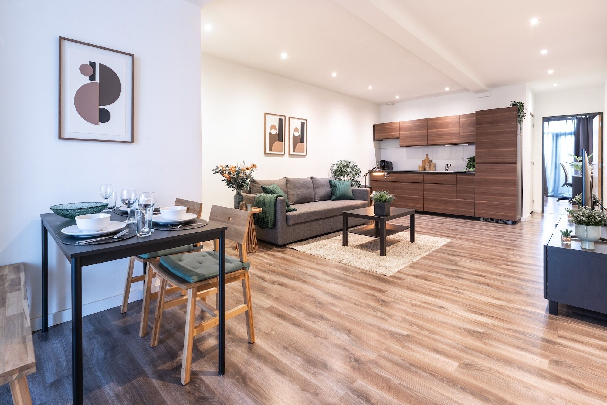 Una sala moderna con comedor, sofá gris, piso de madera y cocina con armarios de madera.