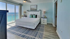 A bedroom with a bed, a ceiling fan, a nightstand, a lamp, a painting, and a view of the ocean.