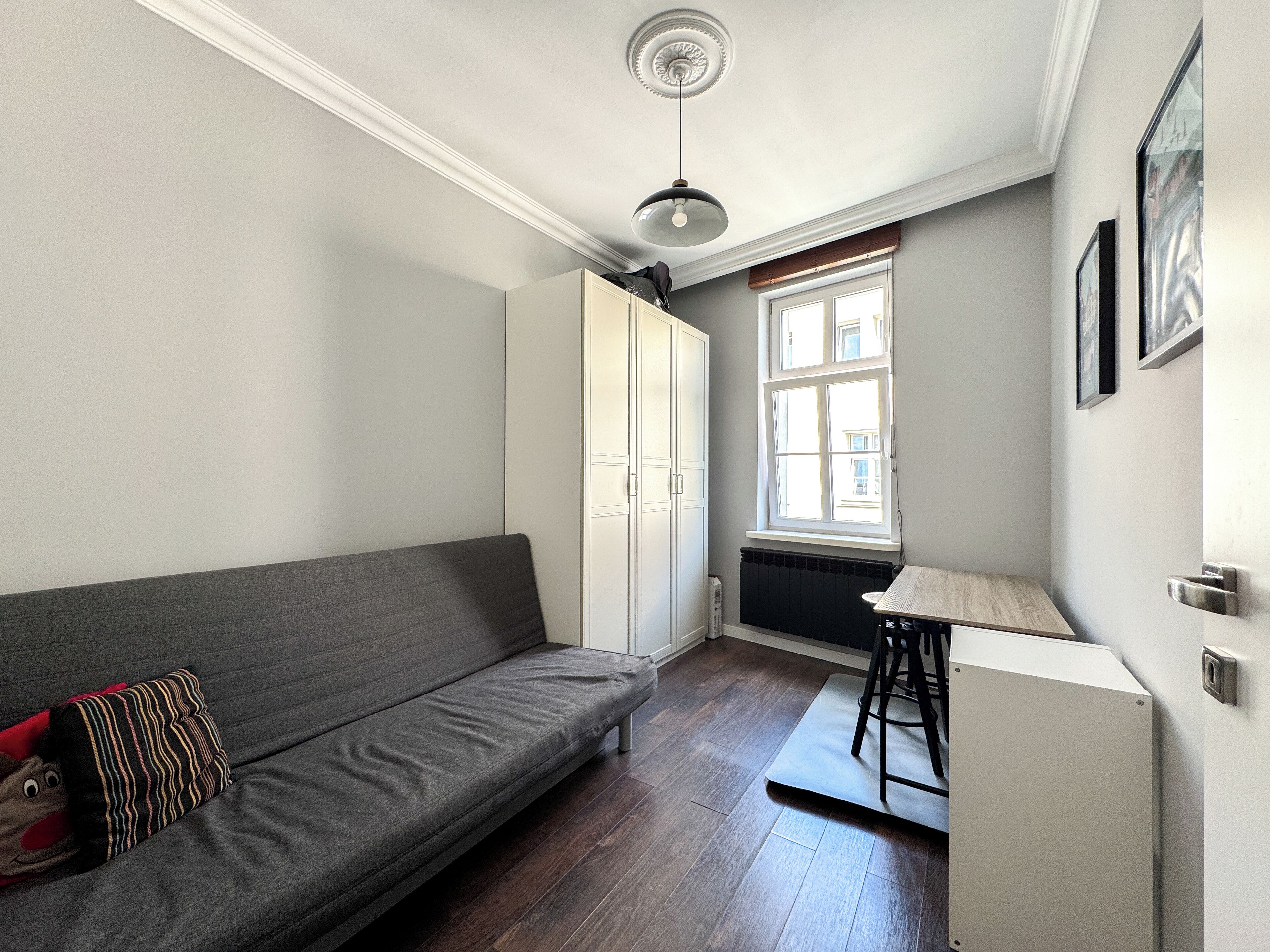 Un espace de vie compact comprenant un canapé gris, une petite table et une fenêtre laissant entrer la lumière naturelle.