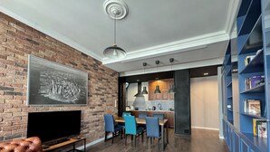 Ett modernt vardagsrum med en tegelvägg, en stor platt-TV, en lädersoffa, en matplats med blå stolar och en bokhylla.