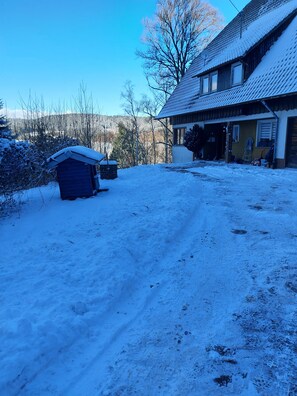 A snow-covered yard with a wooden shed, a house with a sloped roof, and bare trees in the background. - Holiday Apartment with Wi-Fi (Lauterbach)