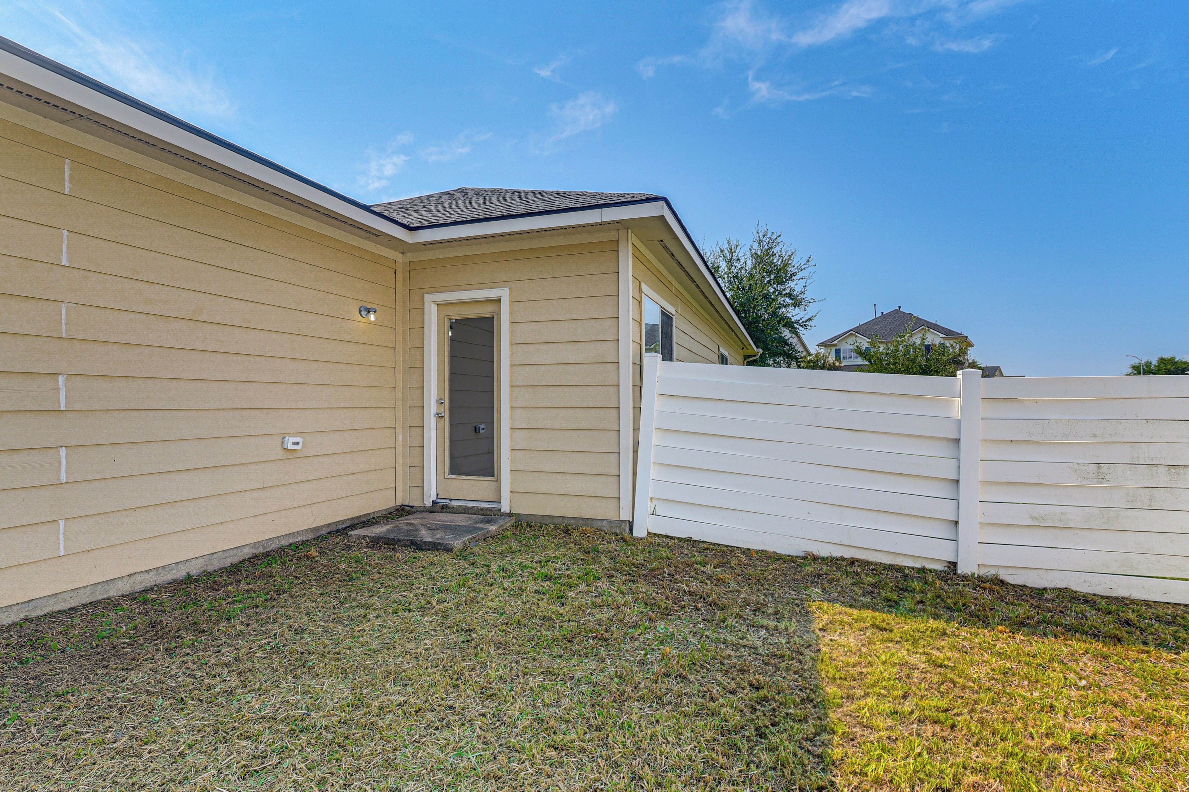 A single-story house with a beige exterior, a front door, and a small front yard with grass and a white fence.