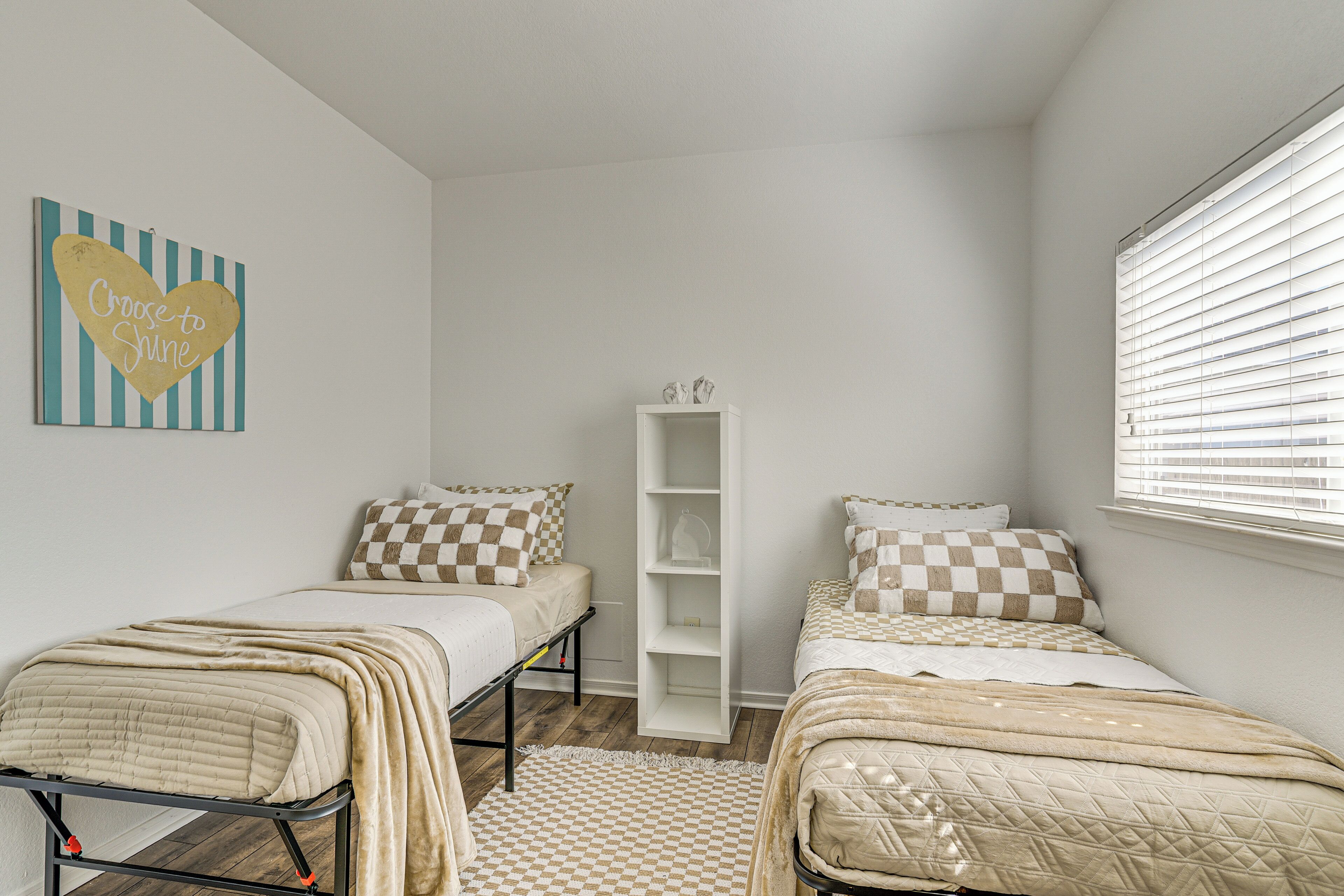 A bedroom with two beds, a checkered rug, a white shelving unit, and a window with blinds.