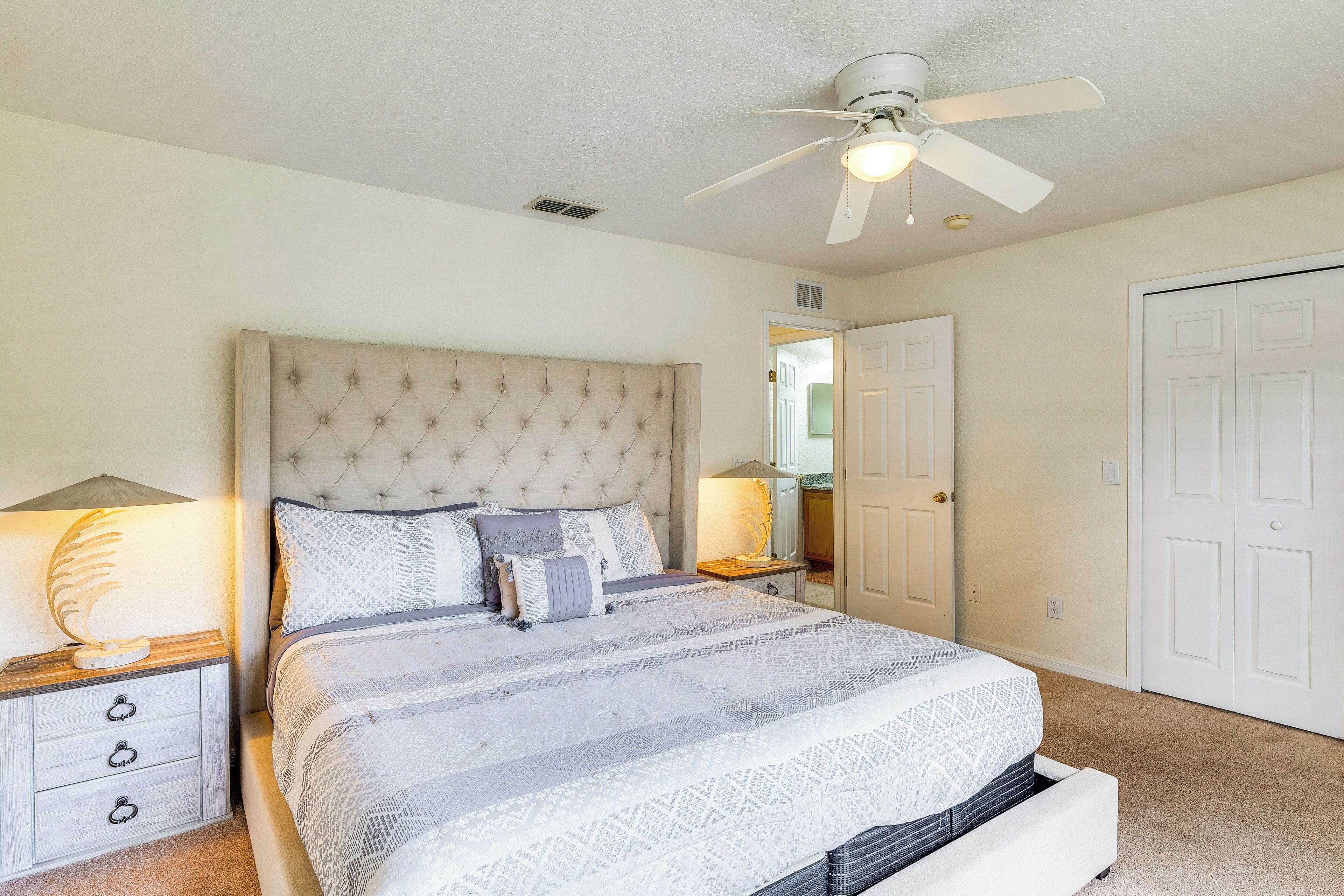 A bedroom with a tufted headboard, a bed with patterned bedding, bedside lamps, a ceiling fan, and a door.