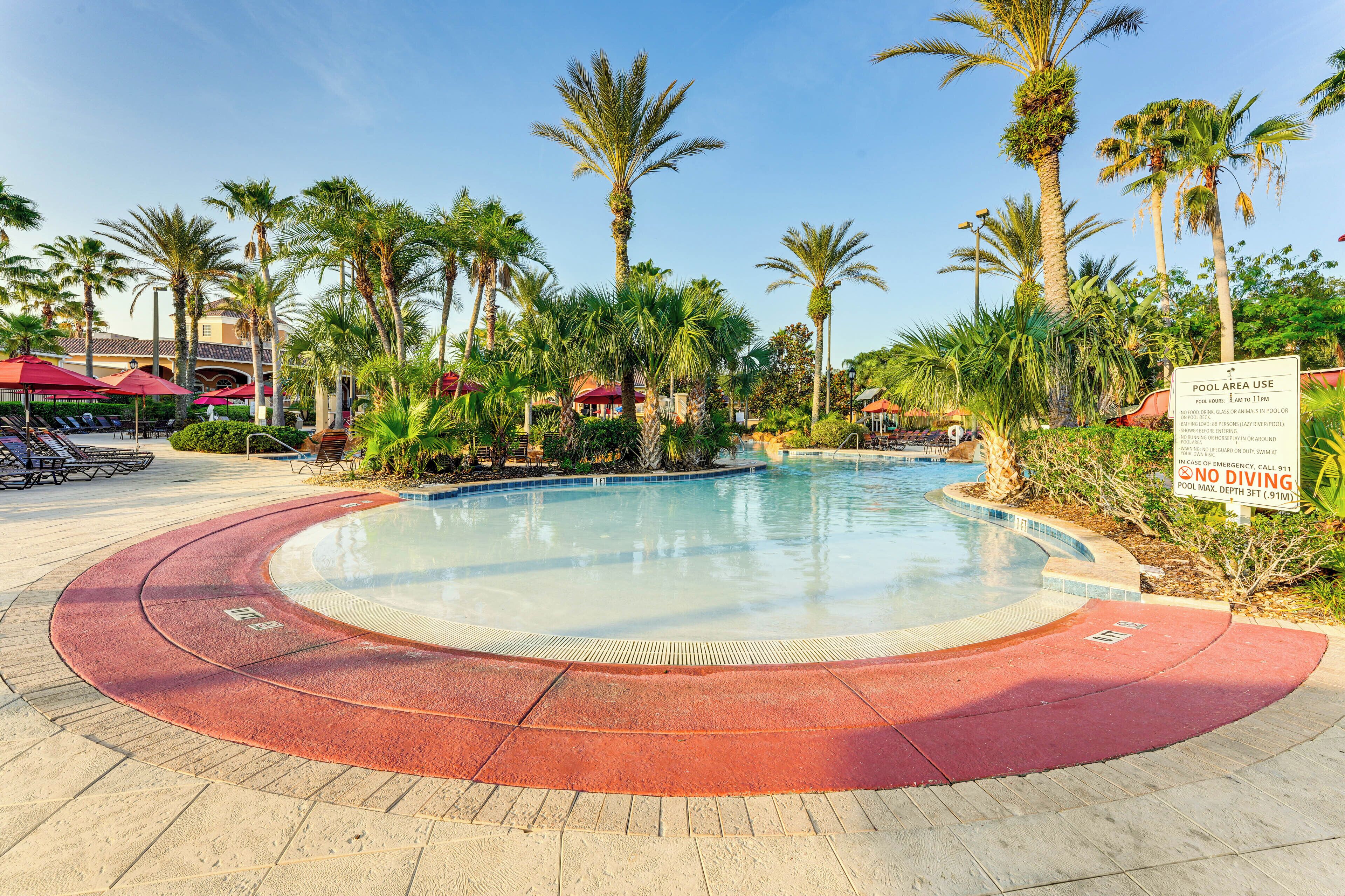 A pool area with a clear blue pool, surrounded by palm trees and red umbrellas.