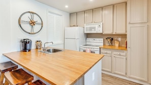 Une cuisine avec un îlot central en bois, des meubles blancs, un four à micro-ondes, un réfrigérateur et une horloge murale.