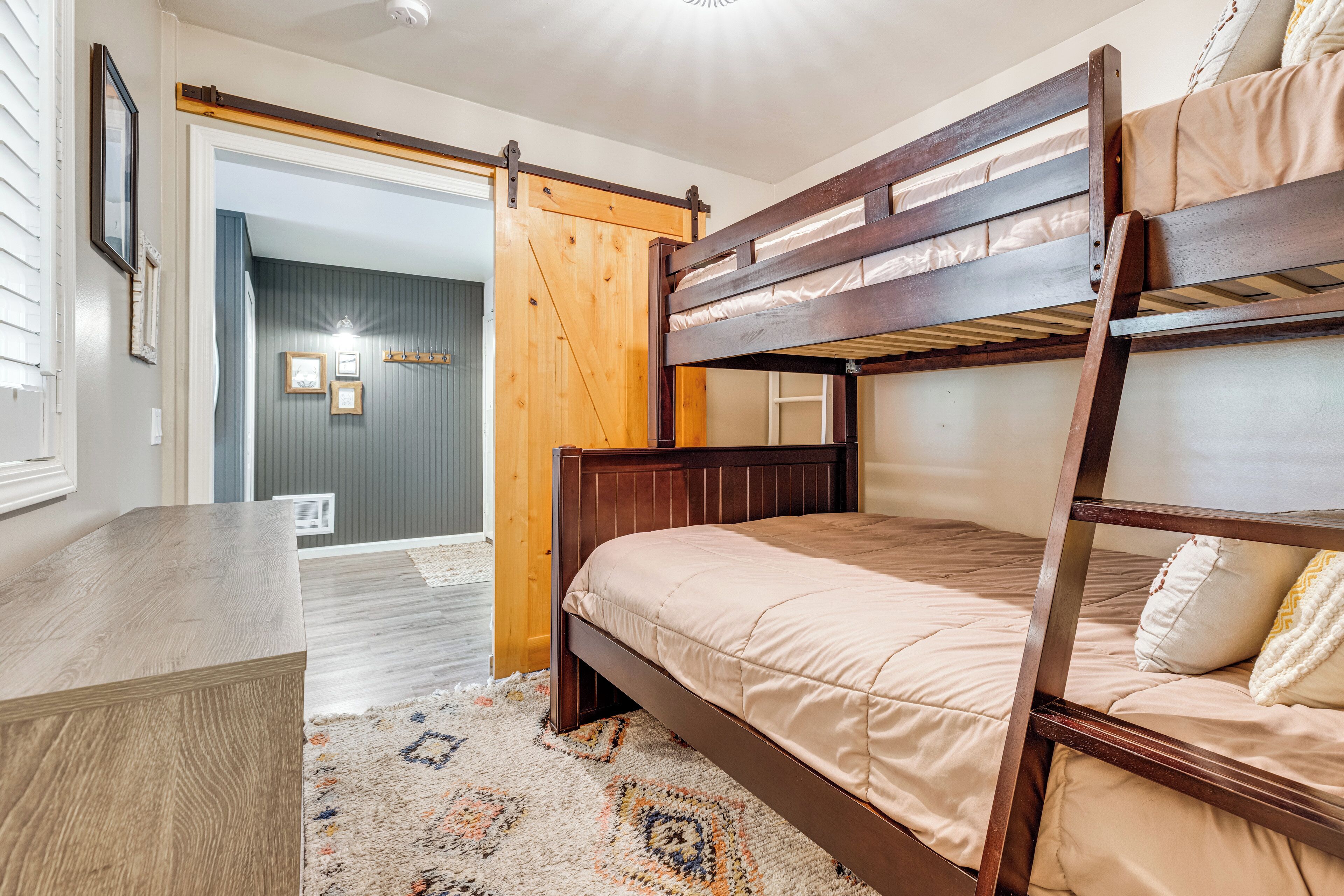 A bunk bed with a wooden frame and beige bedding, a wooden desk, and a patterned rug.