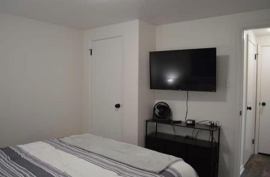 A bedroom with a bed, a TV mounted on the wall, and a small table with a fan.