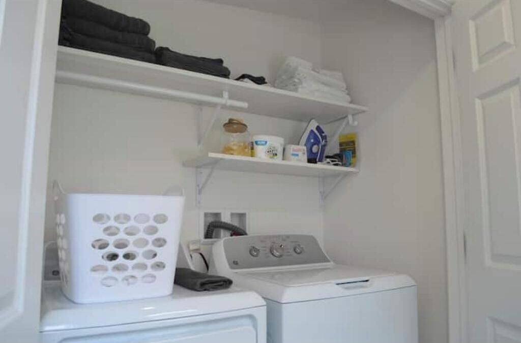 A laundry area with a washing machine, dryer, and shelves above containing folded clothes and laundry supplies.