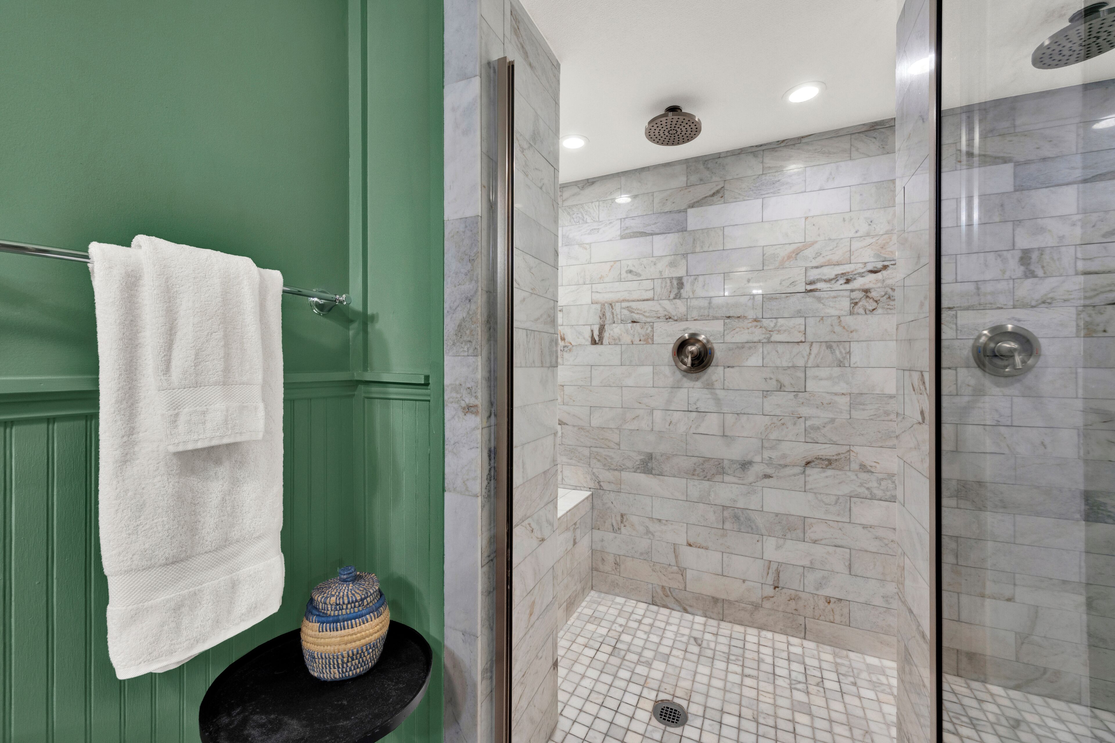 A bathroom with a green wall, a white towel hanging on a rack, a glass-enclosed shower with marble tiles, and a small black sink with a decorative jar.