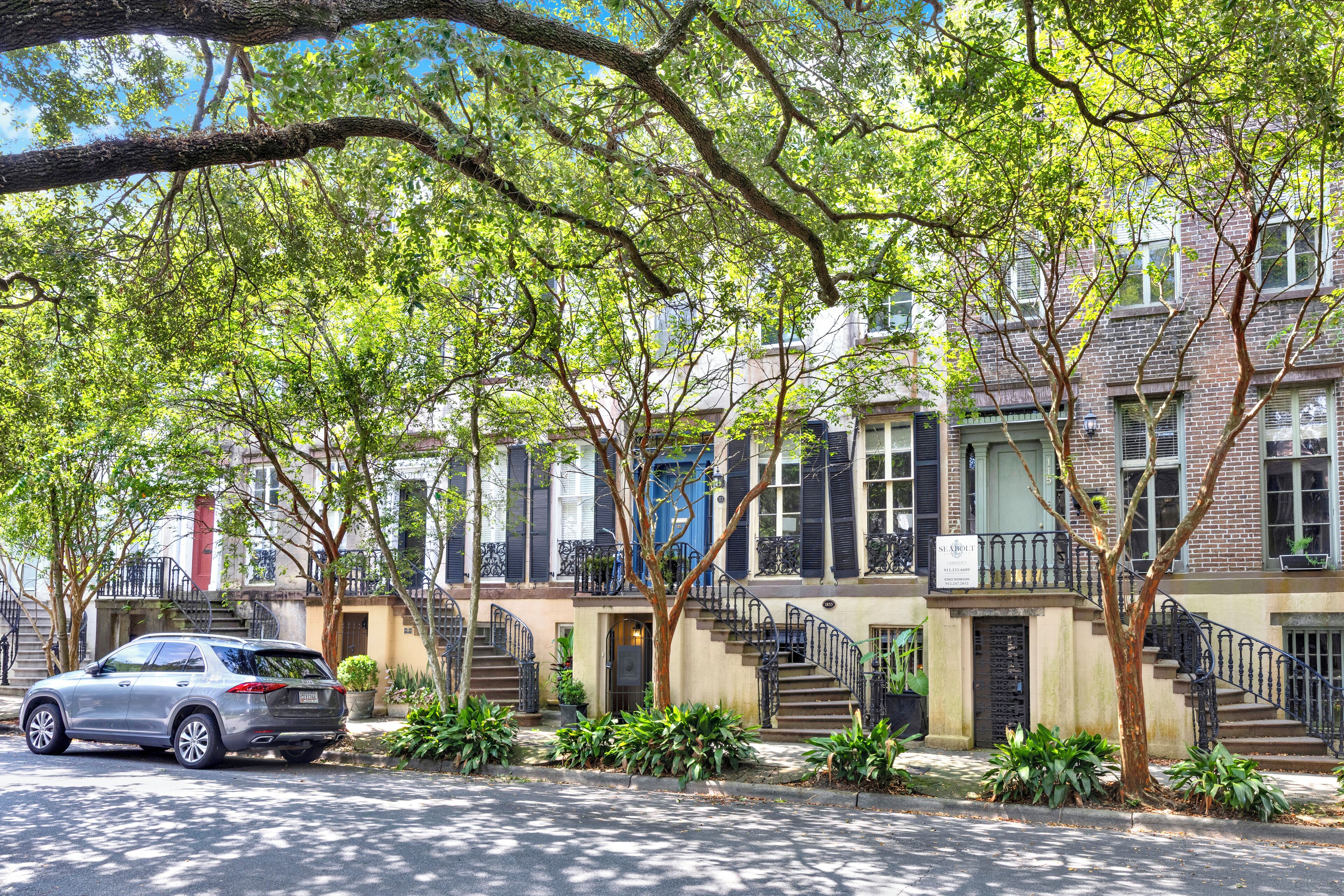 A row of townhouses with a car parked in front, a staircase leading to the entrance, and a tree providing shade.