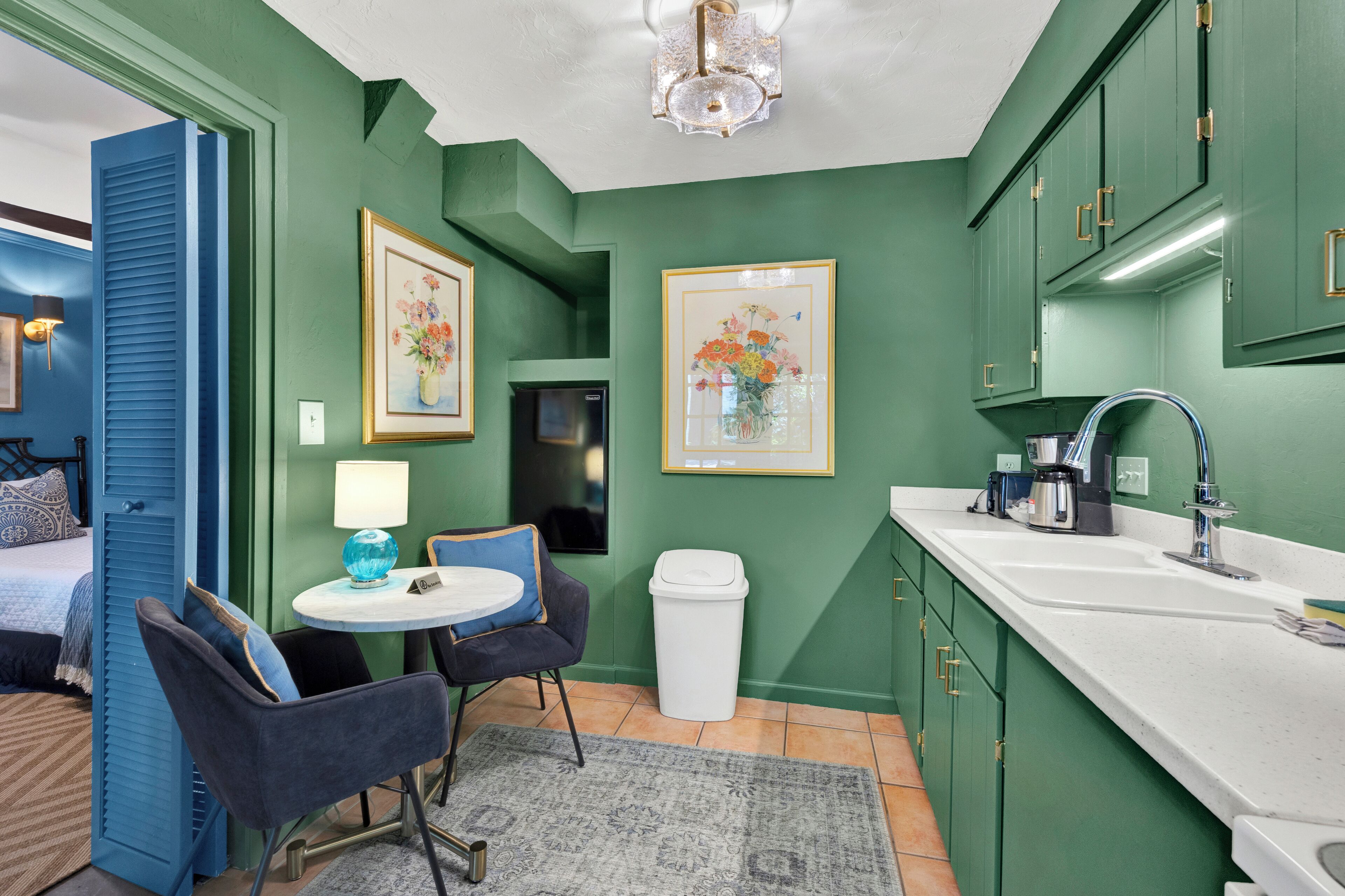 Une cuisine compacte avec des meubles verts, un plan de travail blanc et un évier. On y trouve un petit coin repas avec deux chaises et une table, une poubelle et, au mur, des œuvres d’art florales encadrées.