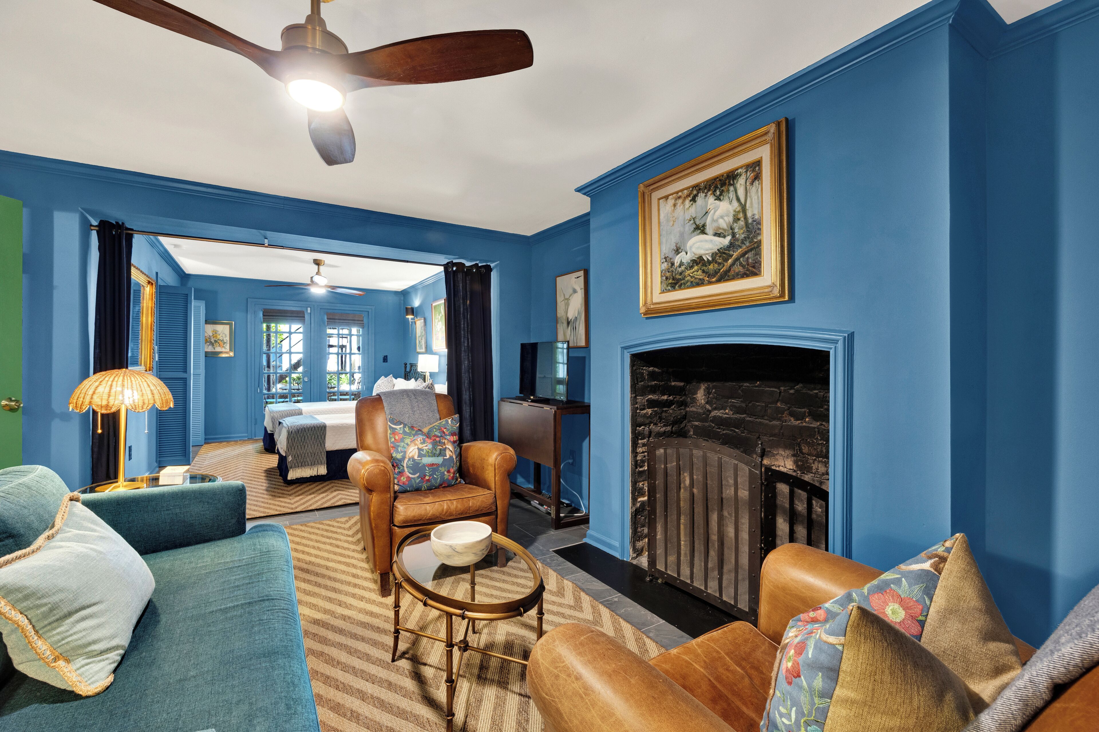 A room with a blue wall, a fireplace, a ceiling fan, and a bed with a view.