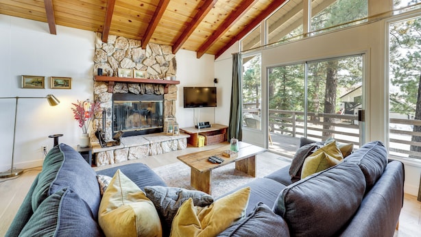 A living room with a stone fireplace, a flat-screen TV, and a large window offering a view of trees. - Game Room + Deck: Wooded Big Bear Lake Retreat (Big Bear Lake)
