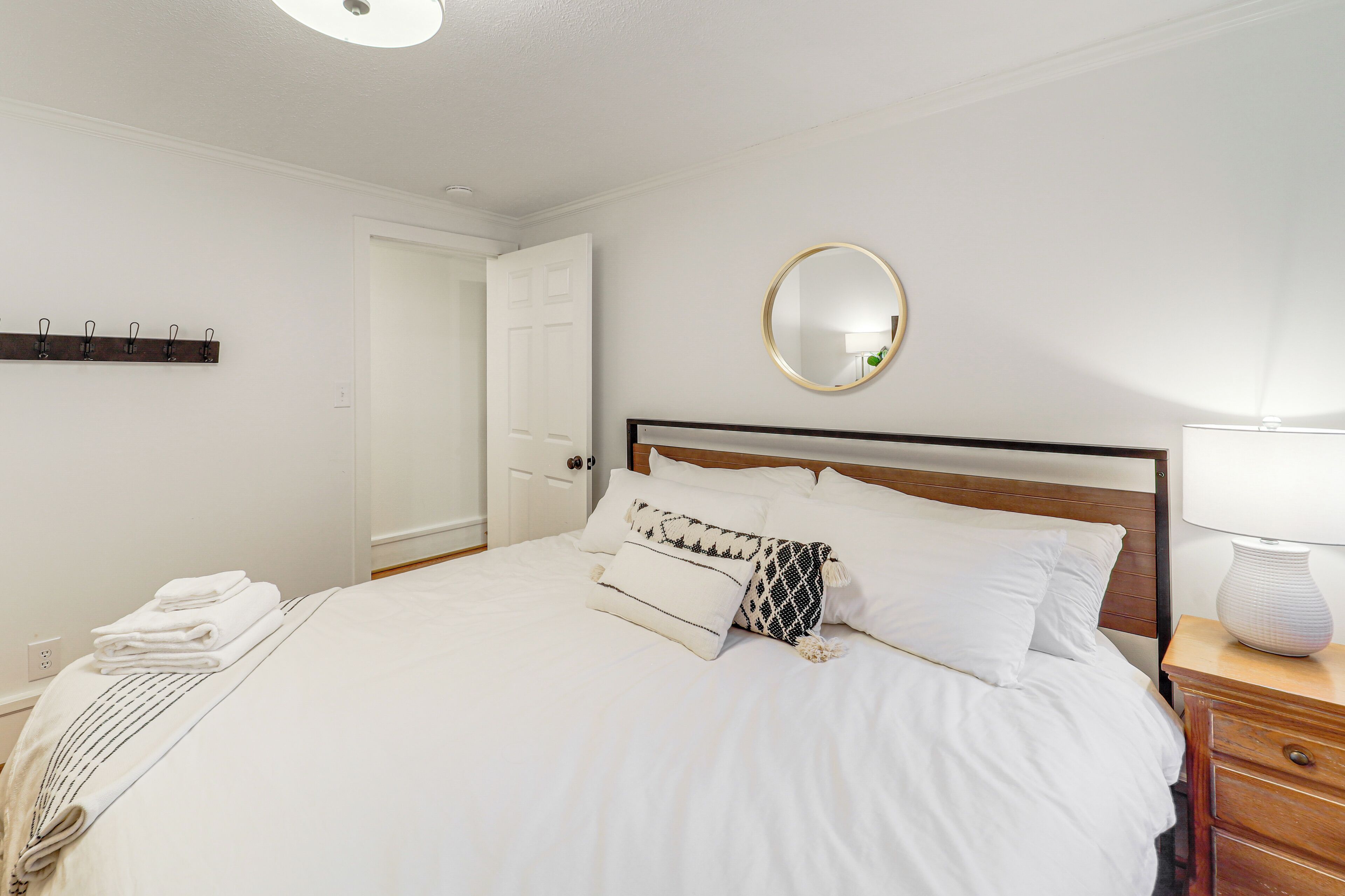 A neatly made bed with white linens and a patterned throw, a bedside table with a lamp, and a wall-mounted mirror.