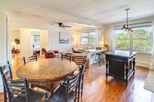 A spacious living room with a wooden dining table, comfortable seating, a fireplace, and large windows offering a view of greenery.