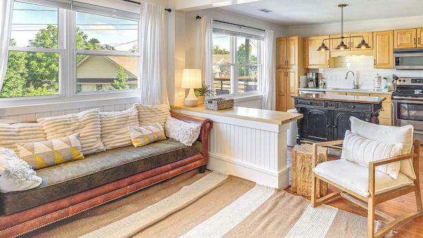 Una sala de estar acogedora con un sofá y un sillón, una zona de cocina con armarios de madera y electrodomésticos, y ventanas grandes con cortinas.