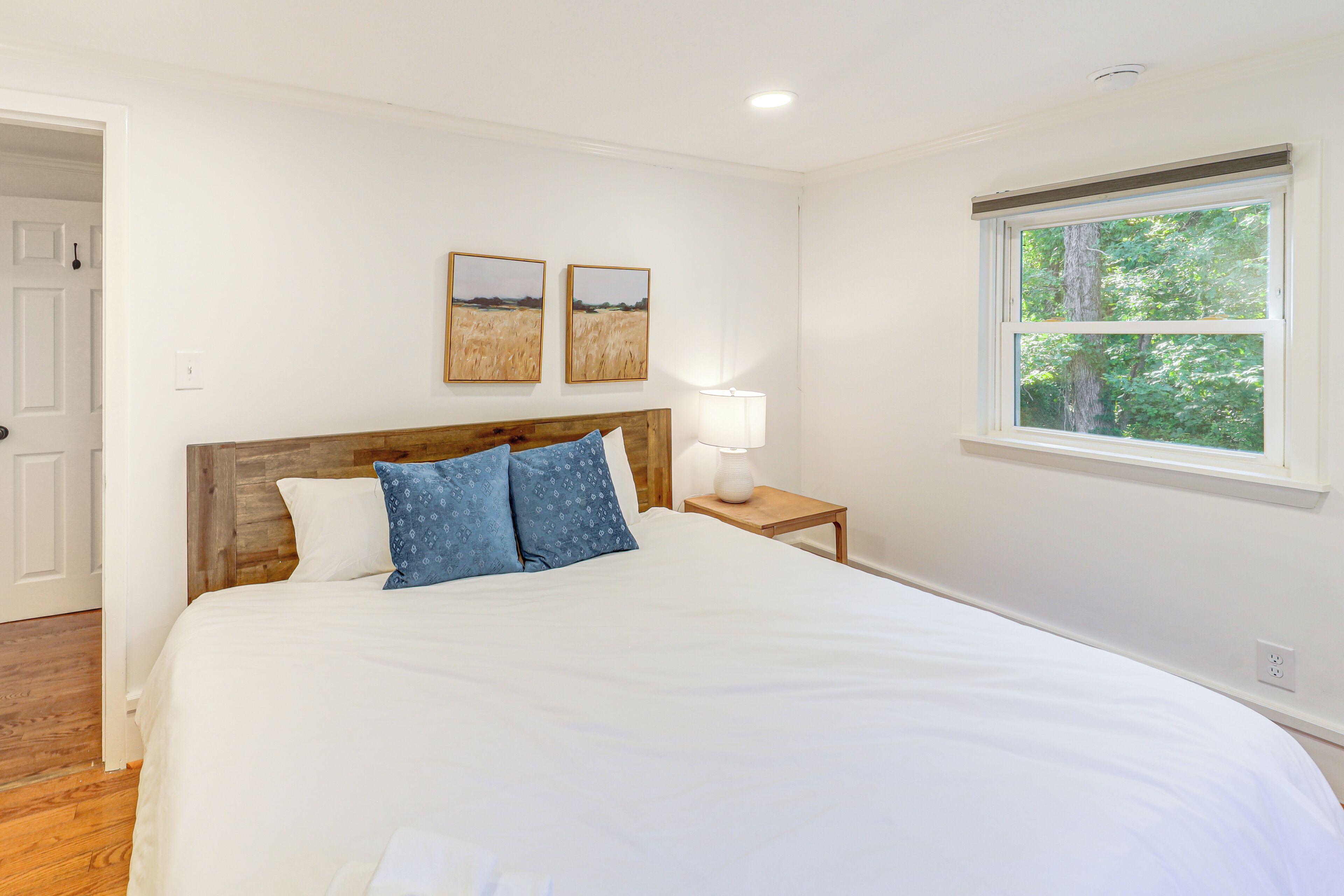 A bedroom with a large bed, a wooden headboard, two wall-mounted paintings, a bedside lamp, and a window with a view of trees.