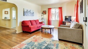 A living room with a red sofa, beige sofa, and a coffee table. There is a TV, a lamp, and a window with curtains. - Las Cruces Home w/ Fenced Yard: Recently Remodeled (Las Cruces)