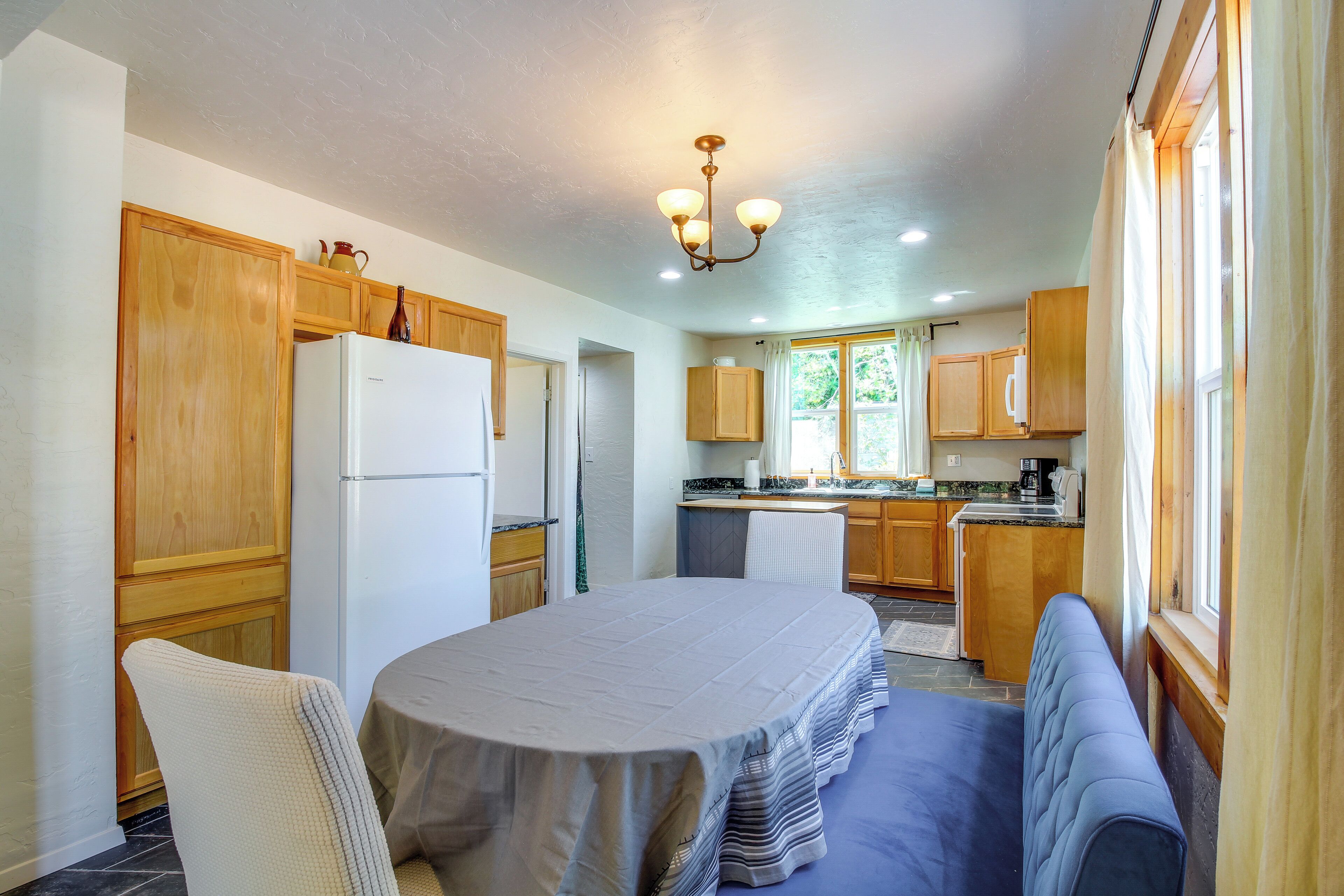 A kitchen with wooden cabinets, a refrigerator, a dining table with a bench, and a window with curtains.