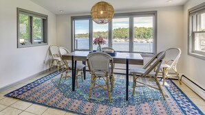 Comedor con una mesa grande, cuatro sillas de mimbre, un tapete estampado y vistas al agua a través de amplios ventanales.