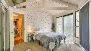 A bedroom with a bed, a nightstand, a lamp, a potted plant, and a view of the outdoors through a large window.