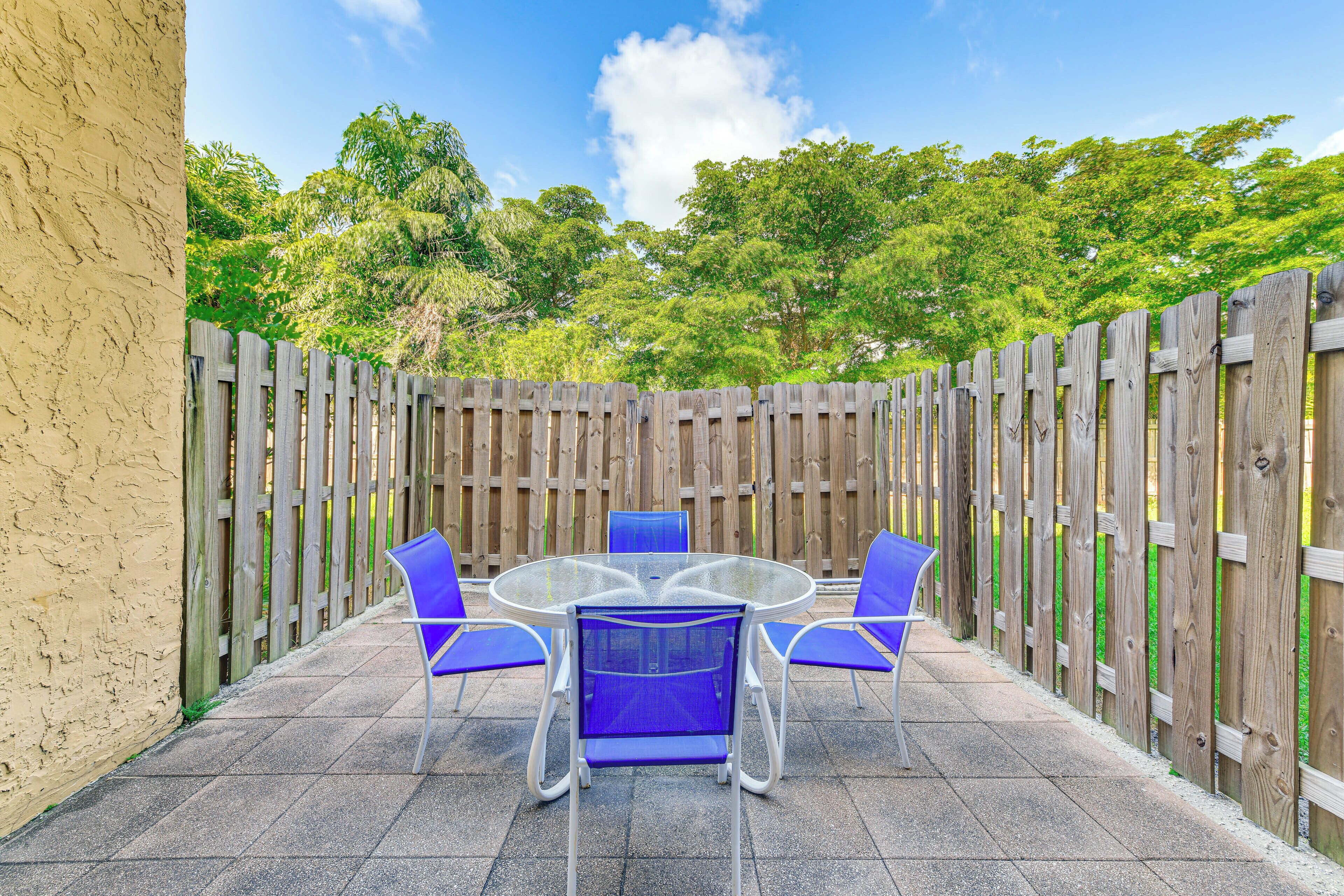 A patio with a round glass table and four blue chairs, surrounded by a wooden fence and a beige wall.