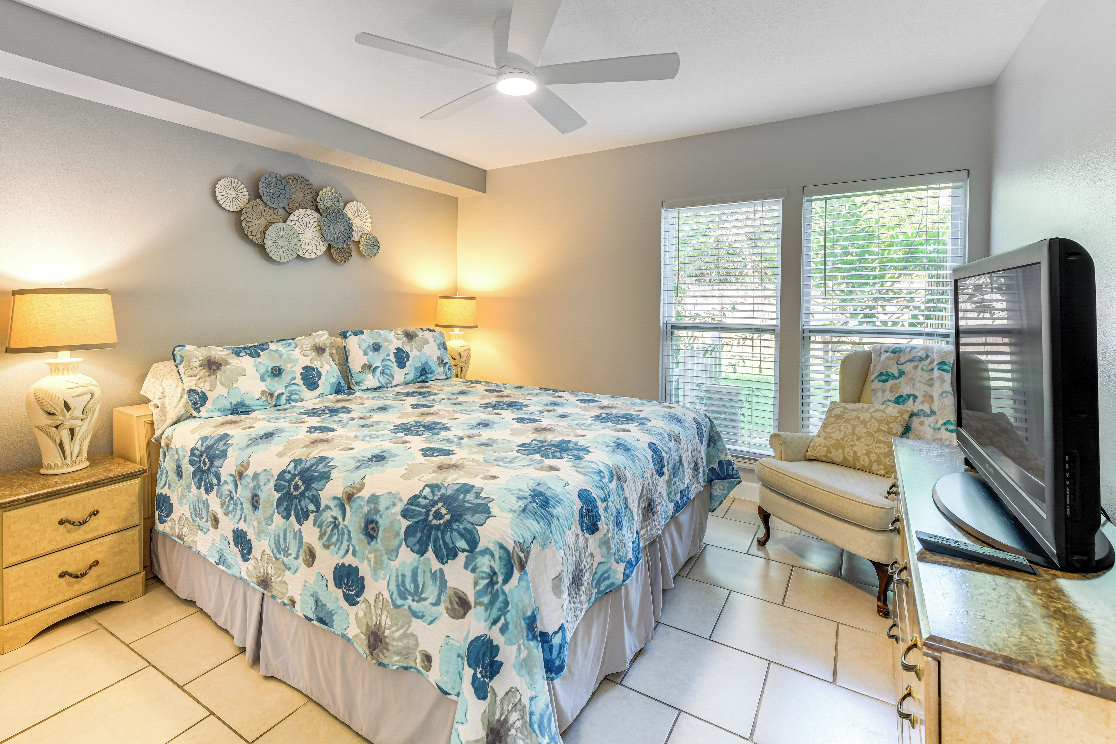 A bedroom with a large bed, a TV, a chair, and a ceiling fan.