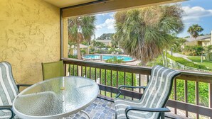 Un balcon doté d’une table en verre et de chaises à rayures, offrant une vue sur une piscine et une végétation luxuriante.