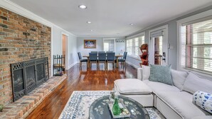 A spacious living room with hardwood floors, a fireplace, a dining area with a table and chairs, and a large window with blinds. - Pool & Deck: Pond-view Getaway in Newnan! (Newnan)