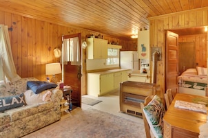 A cozy cabin interior with a wooden floor, a sofa with a 'RELAX' pillow, a dining table, a kitchenette, and a bed with a green and white pillow.