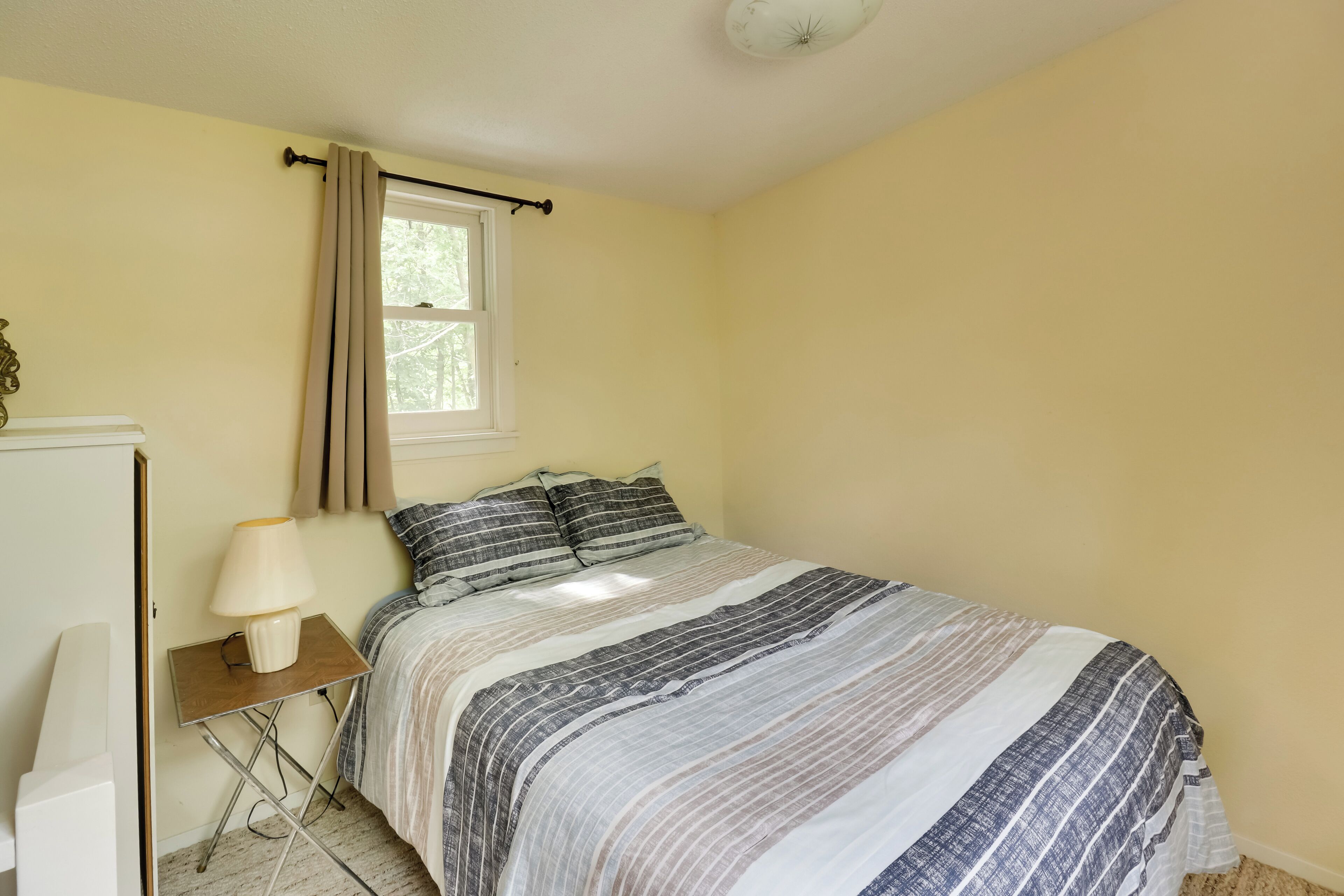 A bedroom with a bed, a bedside table with a lamp, a window with curtains, and a small desk.