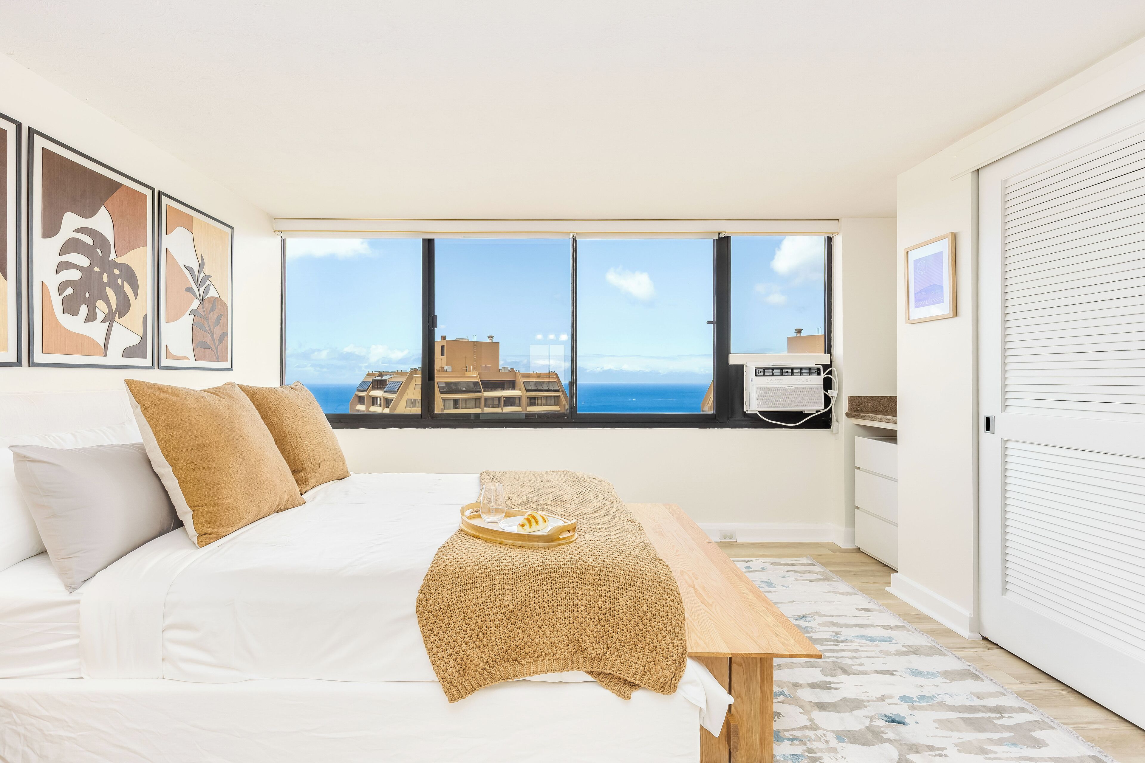 A bedroom with a large window, a bed with white linens, a wooden bedside table, and a view of buildings and the sea.