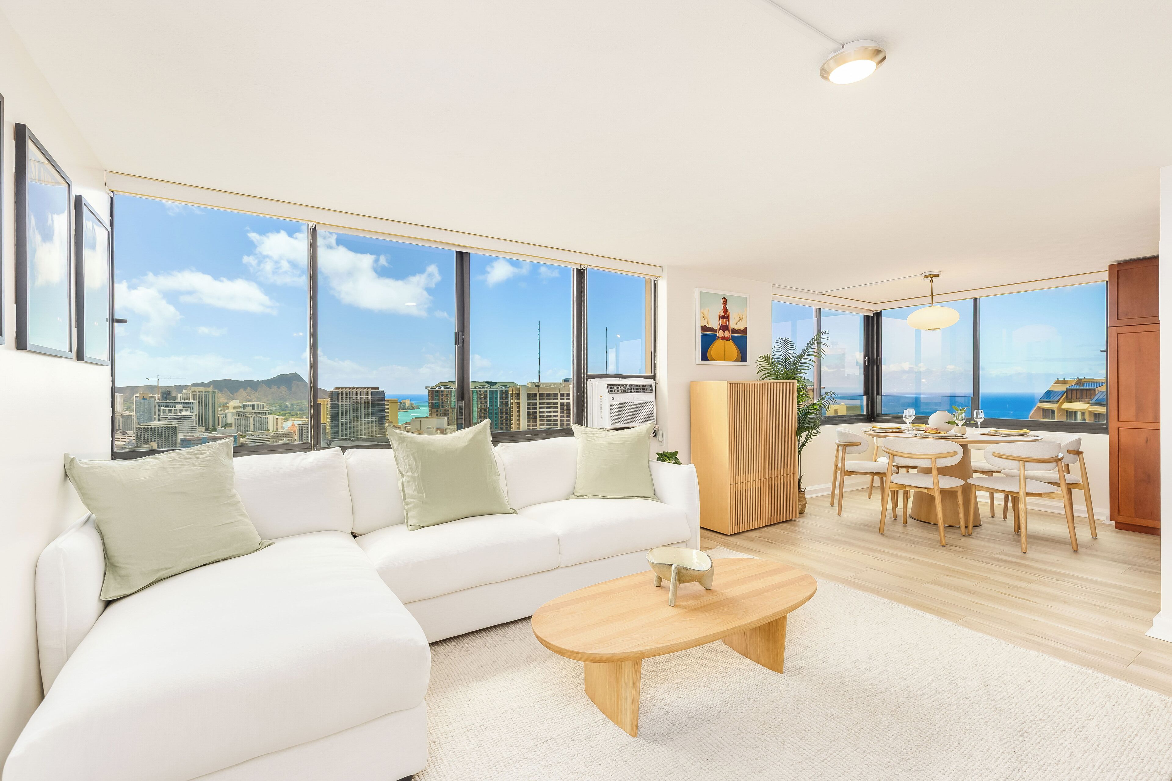 Ein modernes Wohnzimmer mit einem weißen Sofa, einem Holztisch und einem Essbereich mit Holztisch und Stühlen. Große Fenster bieten Blick auf die Stadt.