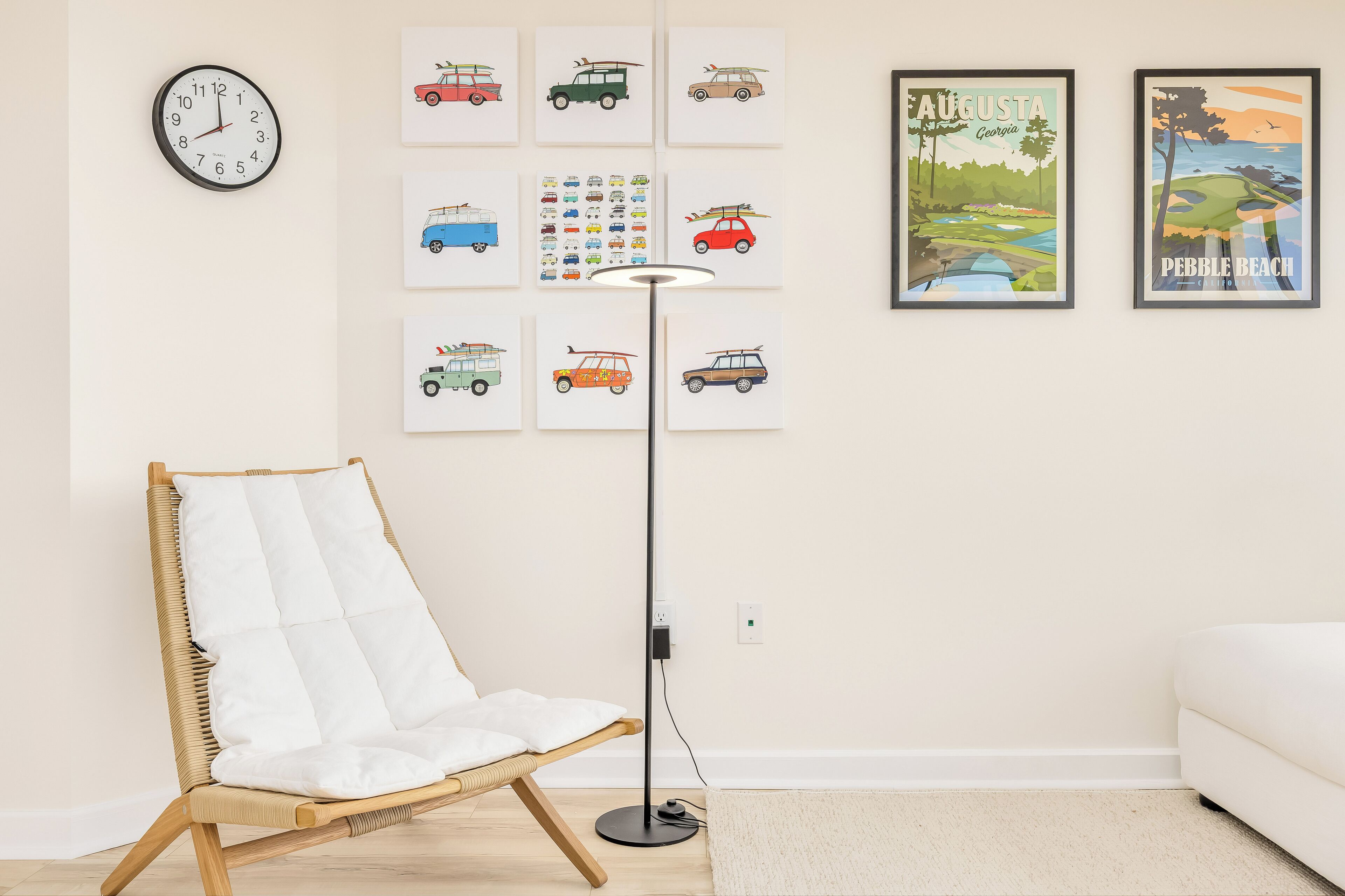 Ein Zimmer mit einem Korbstuhl, einer Stehlampe, einem weißen Sofa und Wandbildern mit Oldtimern und einer Strandansicht.