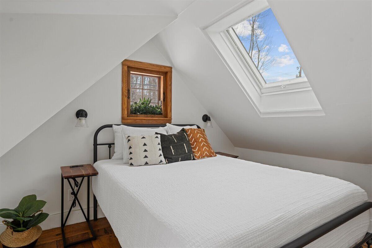 A bedroom with a sloped ceiling, a skylight, a bed with white bedding, and a small bedside table with a plant.