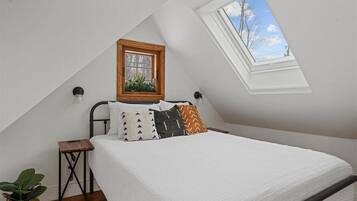 A bedroom with a sloped ceiling, a skylight, a bed with white bedding, and a small bedside table with a plant.