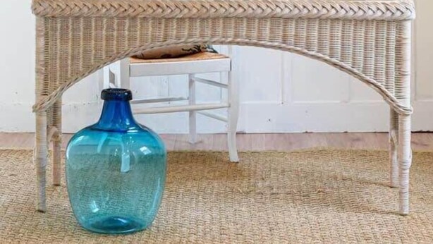 Une pièce avec une table en osier, un vase bleu, une chaise blanche, un luminaire suspendu et une fenêtre donnant sur un espace verdoyant.
