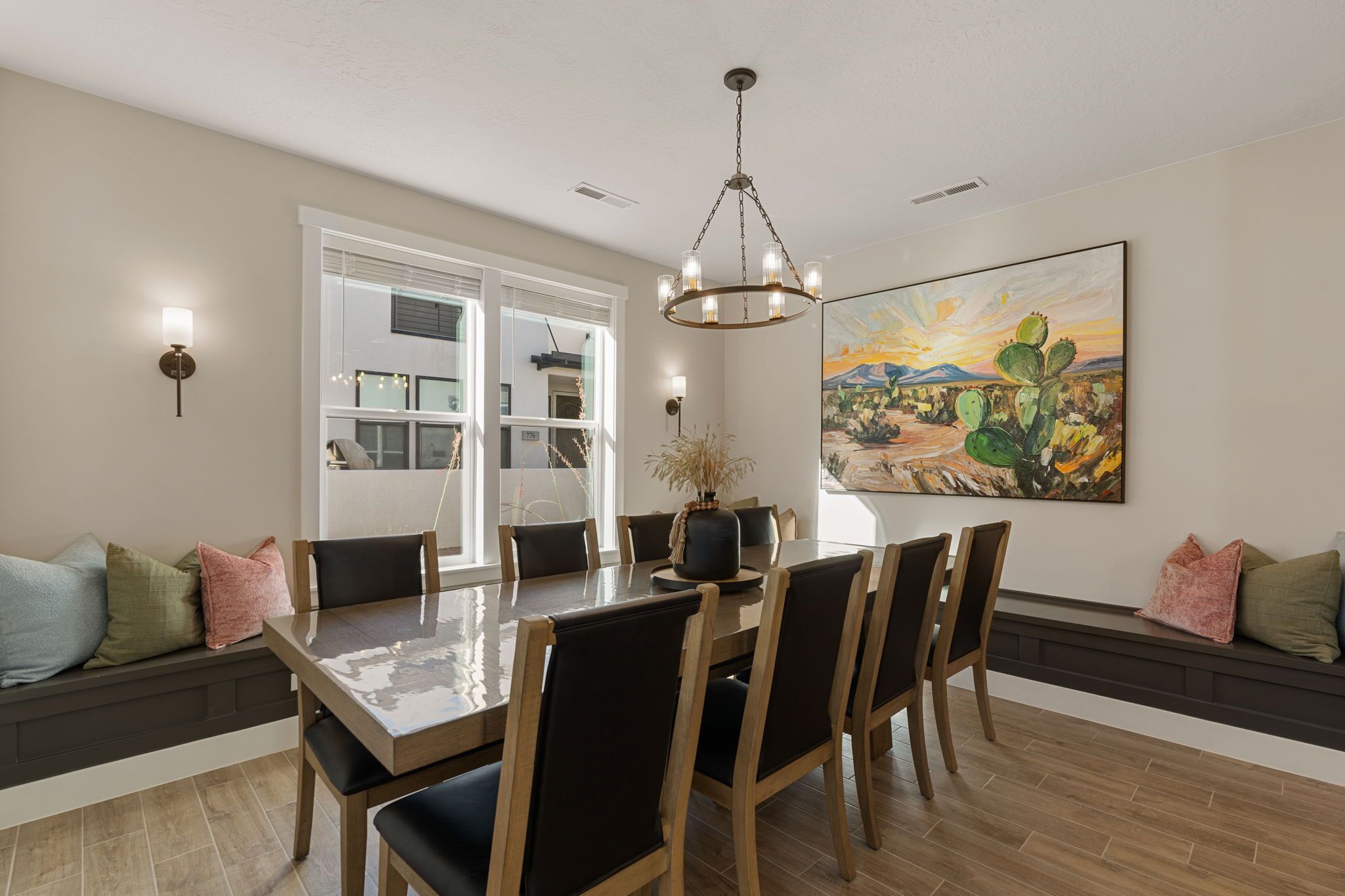 A dining area with a large table, chairs, and a chandelier. There is a painting on the wall and a window with blinds.