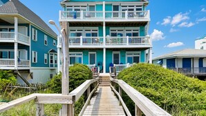 A multi-story building with a wooden deck leading to the entrance, surrounded by greenery. - Octopus's Garden | Oceanfront Retreat w/ Elevator + Beach Access (Carolina Beach)
