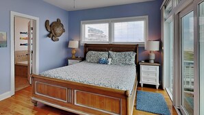 A bedroom with a wooden bed, two bedside tables, a window with blinds, and a view of the outside. - Octopus's Garden | Oceanfront Retreat w/ Elevator + Beach Access (Carolina Beach)