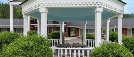 A gazebo with a white exterior and a green roof, surrounded by greenery and a white fence.