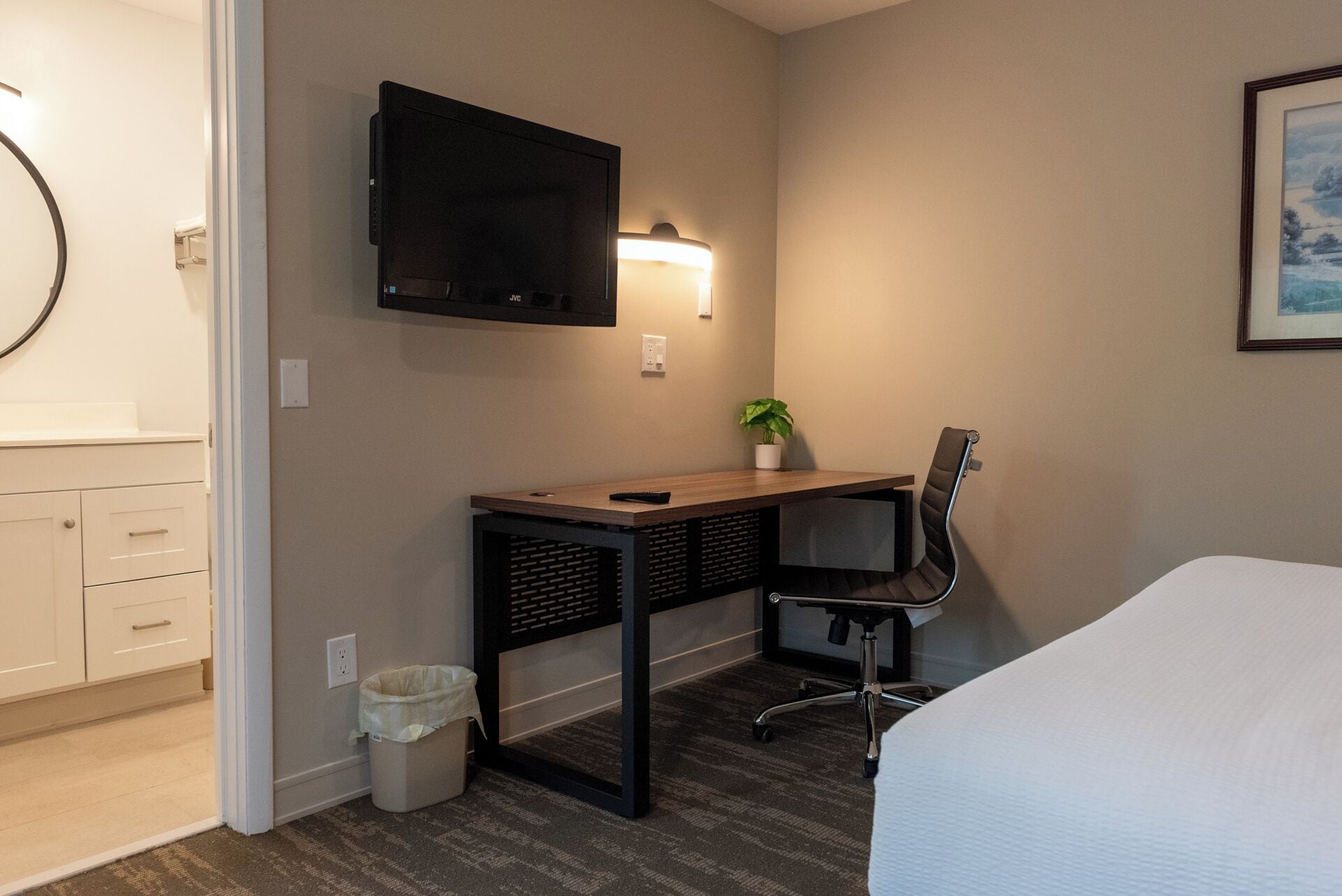 Ein Hotelzimmer mit einem Bett, einem Schreibtisch mit Stuhl, einem an der Wand befestigten Fernseher, einem kleinen Topfpflanzen und einem Mülleimer.