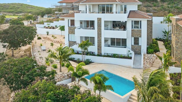 Une maison moderne à plusieurs étages, dotée d’une piscine, entourée d’une végétation luxuriante et située dans un paysage vallonné.