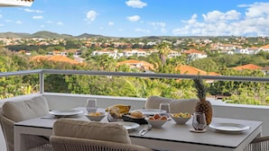 Un balcon avec une table préparée pour le petit-déjeuner, donnant sur un quartier résidentiel avec des maisons et de la verdure.