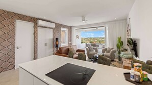 Une cuisine moderne dotée d’un îlot central, d’un coin repas avec un canapé et des fauteuils, et d’une grande fenêtre offrant une vue sur la ville.