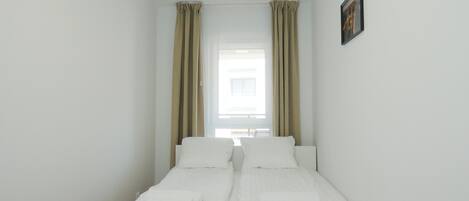 A small, clean room with two single beds, beige curtains, and a framed picture on the wall.