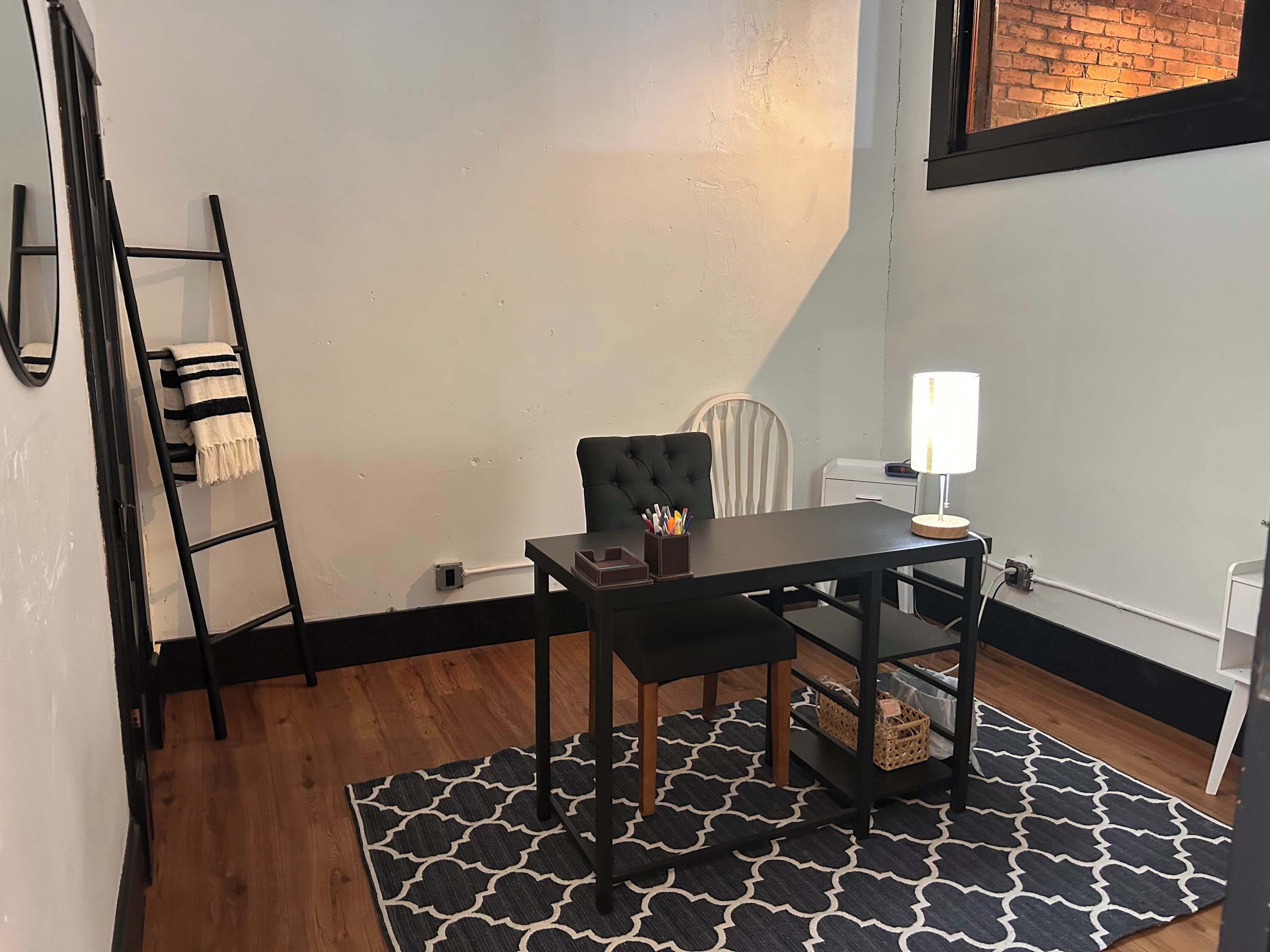 Una habitación pequeña y ordenada, con un tapete de diseño blanco y negro, una silla negra, una mesita con una lámpara y una escalera apoyada en la pared.